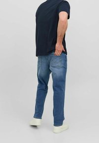 Vaqueros de mezclilla azul con corte recto, lavado más claro en la parte trasera, dos bolsillos traseros y diseño sin costuras, combinados con una camiseta oscura.