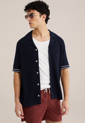 Mees, kes kannab tume sinist lühikese varrukaga nööbitavat kardigani valge t-särgi, burgundiapunaste lühikeste pükste ja päikeseprillidega, vaatab vasakule ühevärvilise tausta ees.