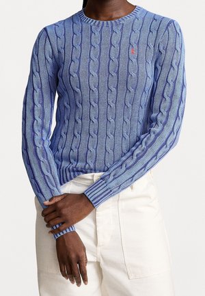 Personne portant un pull torsadé bleu clair avec un petit logo rouge sur la poitrine et un pantalon blanc cassé, les mains croisées à la taille.
