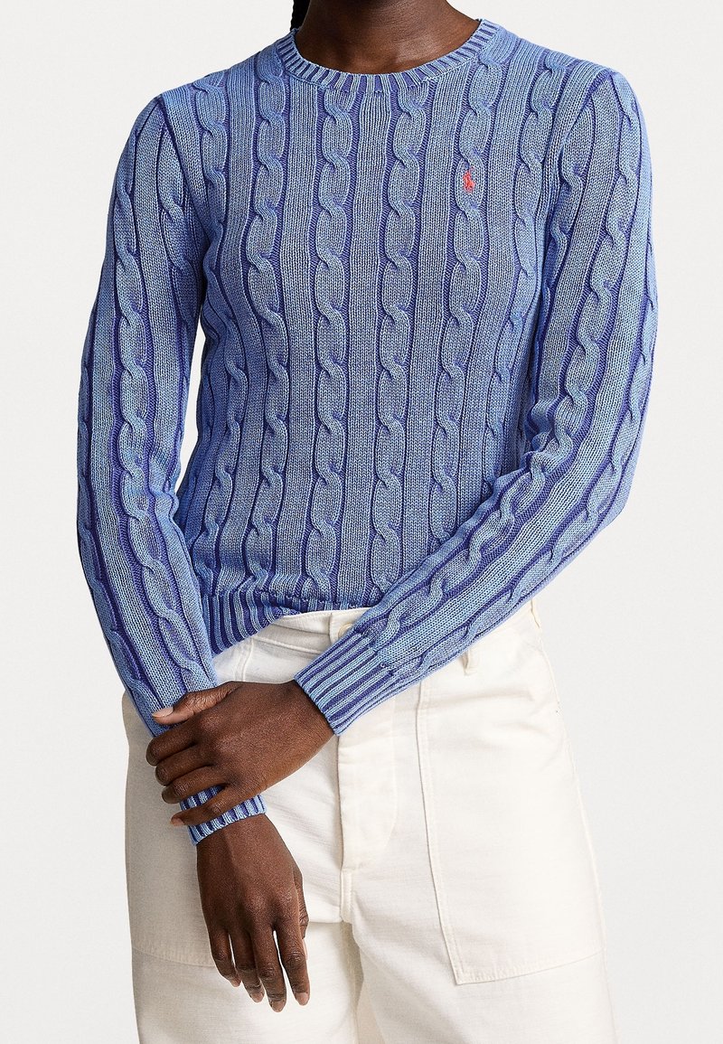 Personne portant un pull torsadé bleu clair avec un petit logo rouge sur la poitrine et un pantalon blanc cassé, les mains croisées à la taille.