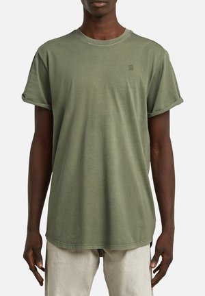 Uomo con maglietta verde oliva a maniche corte con risvolti abbinata a pantaloni beige chiaro, in piedi davanti a uno sfondo semplice.