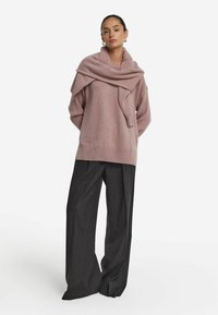 Übergroßer rosa Strickpullover mit umgeschlagenem Kragen und geripptem Saum, kombiniert mit dunkelgrauen, weit geschnittenen Hosen, die eine weiche Textur aufweisen.