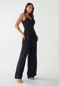 Black crinkled jumpsuit with thin straps, fitted bodice, wide legs, and textured fabric. Paired with light beige strappy heels.