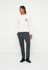 Pullover bianco lavorato a maglia con scollo rotondo, caratterizzato da un dettaglio logo nero. Pantaloni grigi con vita regolabile, abbinati a scarpe eleganti nere.