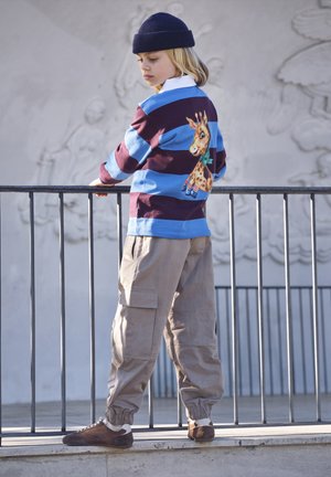 Kind draagt een blauw en bordeaux gestreept shirt met girafprint, beige cargobroek, bruine schoenen en een donkerblauwe muts, leunend op een metalen reling buiten.