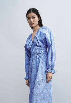 Ung kvinde med langt mørkt hår iført en løs blå kjole med lodrette striber, indsnøret talje, dyb V-halsudskæring og flæseærmer, stående mod en grå baggrund.