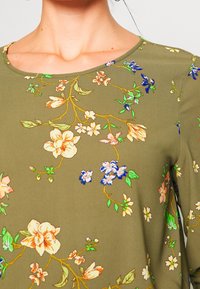 Oliwkowa bluzka wykonana z gładkiego materiału, z okrągłym dekoltem i kwiatowym wzorem z wielokolorowymi kwiatami oraz zielonymi liśćmi.