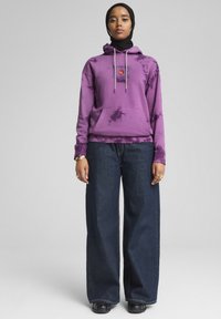 Ljubičasta hoodie sa tie-dye uzorkom, s prednjim džepom i vezicom, u kombinaciji s širokim tamnoplave traperice, prikazuje ležeran izgled.