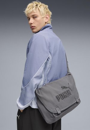 Bolso bandolera gris con solapa, con un logotipo negro de Puma. El modelo lleva una chaqueta azul claro con detalles de malla y pantalones negros.