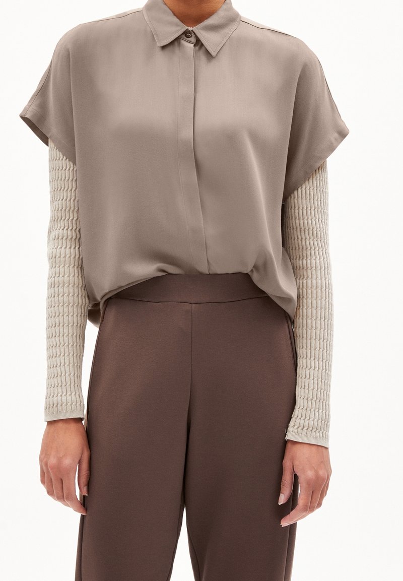 Kortärmad, taupefärgad blus med knappslå framtill; kombinerad med bruna, vida byxor och en texturerad, ljusbeige undertröja med långa ärmar.