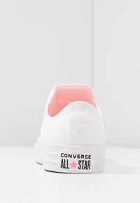 Białe płócienne trampki z różowym wnętrzem i czarną metką z napisem "CONVERSE ALL STAR", posiadające niski krój i gumową podeszwę.