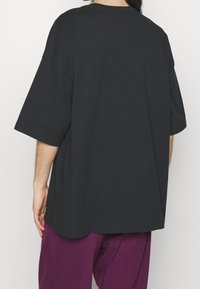 Černé oversized tričko s krátkými rukávy, vyrobené z měkké bavlny. Má kulatý výstřih a zaoblený spodní lem, doplněné volnými fialovými kalhotami.