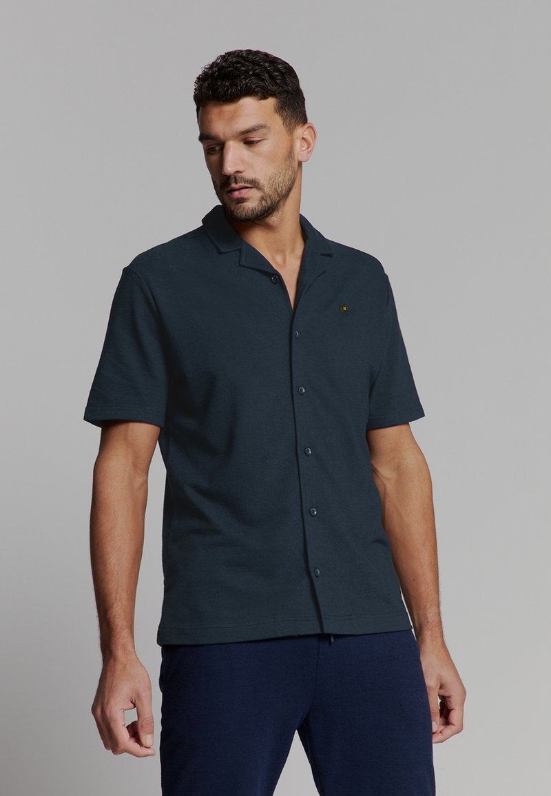 Man met kort krullend haar, gekleed in een donkerblauw overhemd met korte mouwen en marineblauwe broek, kijkt naar beneden naar zijn linkerkant.