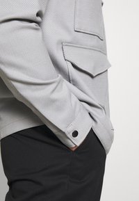 Widok z prawej strony osoby noszącej jasnoszarą, teksturowaną kurtkę z klapkową kieszenią oraz czarne spodnie, ręka swobodnie w kieszeni.