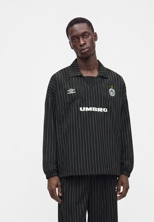 Млад мъж, носещ черна риза Umbro с тънки райета с дълги ръкави и съответстващи панталони, стои на бял фон.