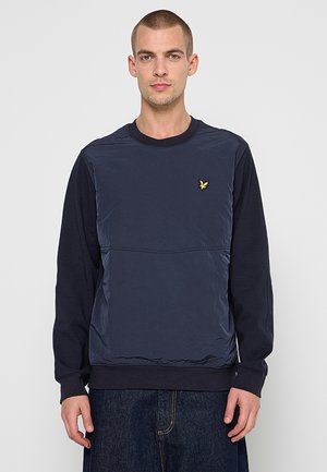 Uomo che indossa una felpa blu navy a maniche lunghe con pannello frontale trapuntato e piccolo logo giallo a forma di uccello sul petto, abbinata a jeans scuri.