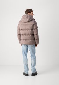 Casaco puffer em padrão xadrez castanho, com capuz, design acolchoado, fecho frontal com zíper e punhos elásticos. Combinado com jeans de lavagem clara e sapatos pretos.