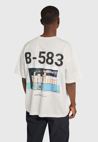 Vit oversized t-shirt med stort svart "B-583" tryck och en färgstark grafisk design på baksidan som visar abstrakta former och färger.