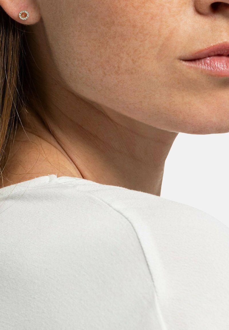 Boucle d'oreille circulaire en or avec une surface texturée, présentée à côté d'un haut de couleur neutre. Teint visible autour de la boucle d'oreille.