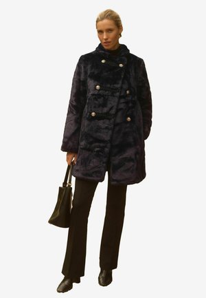 Marineblauer Kunstpelzmantel mit einem doppelt geknöpften Design, glänzenden silbernen Knöpfen, hohem Kragen und seitlichen Taschen, kombiniert mit schwarzen Hosen und einer Handtasche.