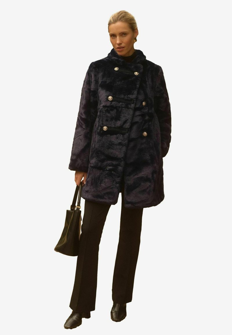 Jūras spēku tumši zila mākslīgās kažokādas mēteli ar dubultu aizdari, spīguļojošām sudraba pogām, augstu apkakli un sānu kabatām, saskaņota ar melnām biksēm un rokassomu.