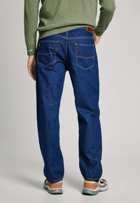 Jeans de mezclilla azul oscuro con un corte relajado, que presentan costuras naranjas y dos bolsillos traseros. Llevados con un suéter verde y zapatillas coloridas.
