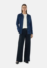 Donkere denim jas met twee borstzakken, gecombineerd met wijdvallende zwarte jeans. Witte top eronder en zwarte puntige schoenen. Gladde textuur.