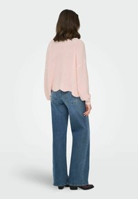 Femme vue de dos, portant un pull en tricot rose clair avec un ourlet festonné et un jean bleu à jambes larges avec des chaussures noires.