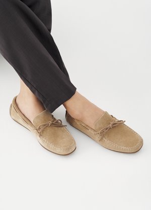 Πόδια με καφέ γούνινες μοκασίνες με διακοσμητικές φιόγκους, συνδυασμένες με σκούρο στενό παντελόνι, πάνω σε λευκό φόντο.