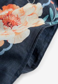 Tissu bleu foncé avec un imprimé de grandes fleurs orange clair et blanches, ainsi que des feuilles vertes, montrant un bord plié et des coutures visibles.