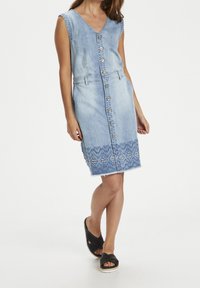 Denimová bezrukávová šaty s výstřihem do V, zapínáním na knoflíky a vyšívaným lemem v bleděmodrém odstínu. Má rovnou siluetu a roztrhané okraje.