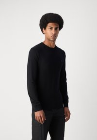 Black long-sleeve sweater made of a smooth knit fabric. Features a crew neckline and a subtle logo on the chest. Worn with dark trousers.