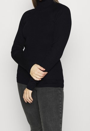 Person i sort ribbet rullekravetrøje og mørkegrå jeans, stående med armene krydset ved håndleddene mod en ensfarvet baggrund.