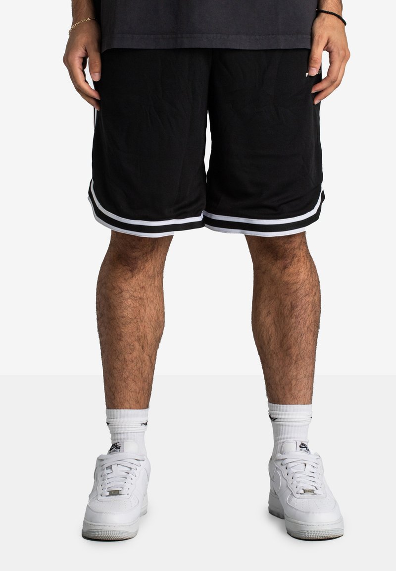 Shorts de sport noirs avec des rayures blanches sur les côtés, fabriqués en tissu respirant. Portés avec des baskets blanches et des chaussettes de sport blanches.