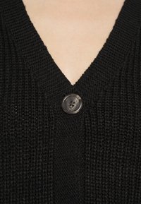 Černý pletený cardigan s výstřihem do V, strukturovanou tkaninou a jedním tmavým mramorovým knoflíkem na zapínání u výstřihu.