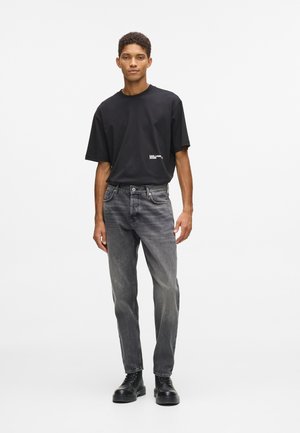 Camiseta negra de manga corta con logo, combinada con jeans grises desgastados y botas negras con cordones. Diseño simple y de corte relajado.