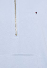 Suéter azul claro con un zipper dorado, que presenta un pequeño logo bordado con franjas rojas, blancas y azules cerca del escote y un bolsillo frontal.