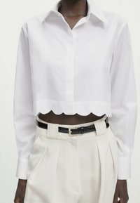 Chemise blanche courte boutonnée avec un ourlet festonné, dotée de manches longues et d'un col classique, associée à un pantalon beige taille haute et une ceinture noire.