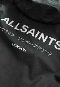 Sort vandtæt jakke med gittertekstur, der har fed hvid tekst "ALLSAINTS" og yderligere tekst på japansk samt "LONDON" i bunden.