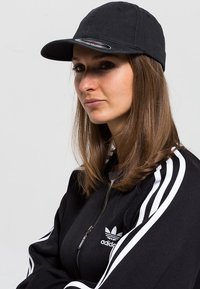 Čierna bavlnená šiltovka s ohnutým šiltom, s logom na prednej strane, nosená s čiernou zipsovou bundou s bielymi pruhmi.