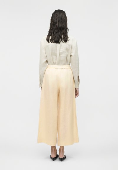 Femeie cu părul întunecat ondulat, purtând o bluză bej cu mâneci lungi, pantaloni largi de culoare piersică deschisă și tocuri negre, stând cu fața spre spate.