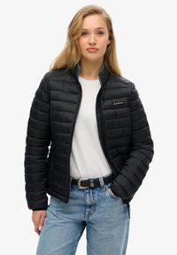 Zwarte gevoerde jas met een ritssluiting aan de voorkant, geborduurd patroon, elastische cuffs en een logo op de borst. Draagt over een wit T-shirt en blauwe jeans.