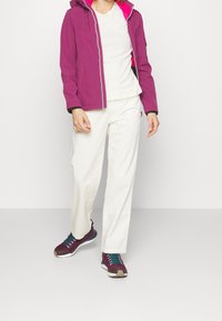 Fioletowa wodoodporna kurtka z różowym wnętrzem, biały t-shirt, kremowe spodnie i bordowe trampki z turkusowymi akcentami, stojące na szarej podłodze.