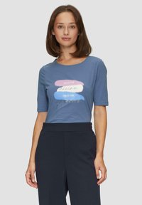 T-shirt bleu à manches courtes avec encolure ronde, arborant un graphisme imprimé en rose, blanc et bleu, avec des accents de texte argenté.