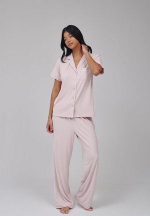 Vrouw staat blootsvoets, draagt een lichtroze kort mouwig pyjamashirt met knopen en losse bijpassende broek, tegen een effen grijze achtergrond.
