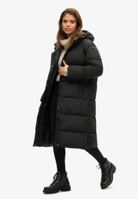 Sort pufferjakke med hætte, med et langt design, kviltet mønster og trykknapper. Bæret over en beige sweater og sorte bukser.