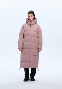 Lang, lichtroze pufferjack met een hoge kraag, horizontale stiksels, een ritssluiting aan de voorkant en zijzakken met rits. Gekocht over zwarte broek en laarzen.