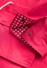 Solapa del bolsillo de la chaqueta roja levantada para mostrar el forro de tela a cuadros rojos y blancos y cierres de botones a presión plateados.