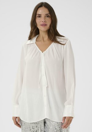 Sieviete valkā baltu blūzi ar garām piedurknēm, pogām un kakla izgriezuma saiti, kā arī sudraba spīguļotās bikses, stāvot pret vienkāršu fonu.