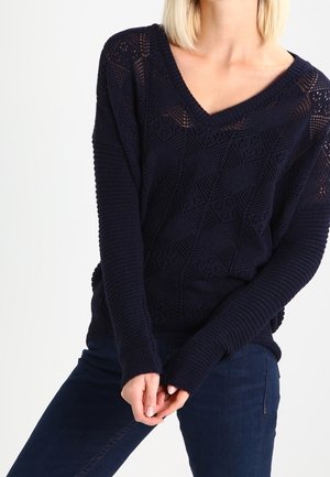 Vrouw draagt een donkerblauw, getextureerd gebreide V-hals trui met lange mouwen en een donkere spijkerbroek, handen voor zich samengevouwen.